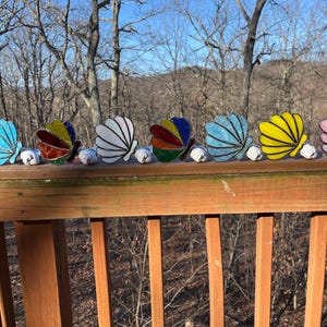 May include: Six stained glass butterfly ornaments in a row, each with a different wing pattern and colour scheme. The butterflies are mounted on small white bases and are displayed on a wooden railing against a backdrop of trees and a blue sky.
