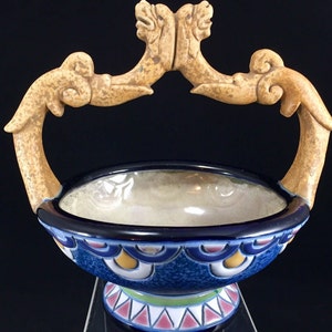 May include: A blue and white ceramic bowl with a decorative handle shaped like two stylised animal heads. The bowl has a blue and white geometric pattern and a yellow and green base.