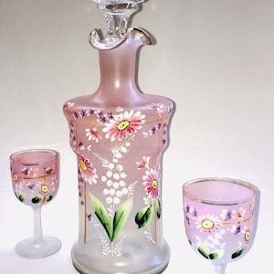 Könnte beinhalten: Ein Set aus drei mattierten Glaskaraffen mit handbemalten Blumendesigns. Die Karaffen sind rosa, weiß und grün. Das Set enthält eine große Karaffe mit einem Stopfen und zwei kleinere Karaffen.