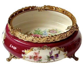 May include: A red and white porcelain bowl with gold trim and floral designs. The bowl has three gold feet and a gold rim.