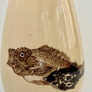 May include: A cream-colored ceramic vase with a brown and black fish design. The vase is shaped like a teardrop and has a smooth, glossy finish.