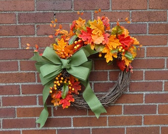 May include: A fall-themed wreath with a grapevine base, adorned with colorful autumn leaves in shades of red, orange, and yellow. A large green bow and sprigs of berries and pinecones add to the seasonal decor.
