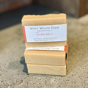 May include: Three rectangular bars of turmeric goat milk soap are stacked. Each bar is wrapped with a white label featuring the text "Wispy Willow Farm" and "Turmeric". The soap is a light beige color.