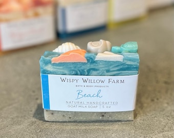 May include: A bar of natural goat milk soap with a beach theme. The soap is blue and white with seashell and turtle decorations. The label reads "Wispy Willow Farm Beach Natural Handcrafted Goat Milk Soap 5 oz."