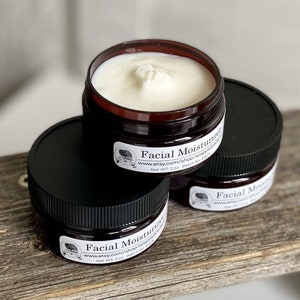 May include: Three small jars of facial moisturizer with black lids and brown bases are stacked on a wooden surface. Each jar has a white label with text and a logo. The jars contain a white cream. The text on the label reads "Facial Moisturizer".