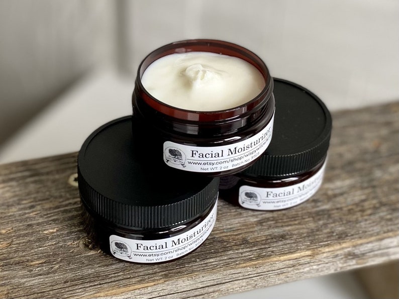 May include: Three small jars of facial moisturizer with black lids and brown bases are stacked on a wooden surface. Each jar has a white label with text and a logo. The jars contain a white cream. The text on the label reads "Facial Moisturizer".