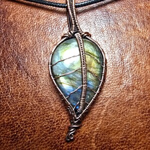 May include: A copper wire wrapped pendant featuring a green and blue labradorite gemstone. The pendant is hanging from a black cord.