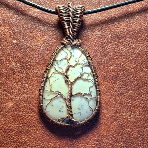 May include: A white and gray dendritic agate stone pendant with a copper wire tree of life design. The pendant is attached to a black cord.