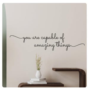 Op de afbeelding: Een witte muur met de tekst "You are capable of amazing things" geschreven in zwarte cursieve letters. Een kleine vaas met witte bloemen staat op een houten plank.