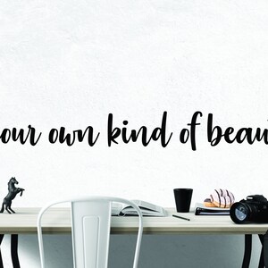 Pode incluir: Uma parede branca com o texto "be your own kind of beautiful" escrito em letra cursiva preta. Uma mesa de madeira com uma cadeira de metal branco, uma caneca preta, um livro, um l&aacute;pis, dois donuts e uma c&acirc;mera preta com uma al&ccedil;a preta est&atilde;o sobre a mesa. Uma pequena figura de cavalo preto est&aacute; sobre a mesa.