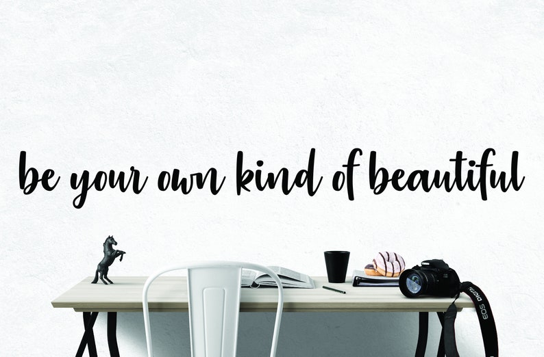 Pode incluir: Uma parede branca com o texto "be your own kind of beautiful" escrito em letra cursiva preta. Uma mesa de madeira com uma cadeira de metal branco, uma caneca preta, um livro, um l&aacute;pis, dois donuts e uma c&acirc;mera preta com uma al&ccedil;a preta est&atilde;o sobre a mesa. Uma pequena figura de cavalo preto est&aacute; sobre a mesa.