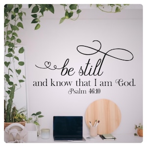 Puede incluir: Una pared blanca con una calcomanía de vinilo negro que dice "be still and know that I am God. Psalm 46:10". La calcomanía está sobre un escritorio con una computadora portátil, una tabla de cortar de madera y un frasco de vidrio con lápices.