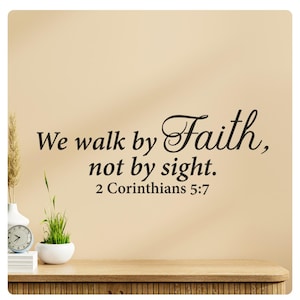 Puede incluir: Una pared beige con una calcomanía de pared de vinilo negro que dice "We walk by Faith, not by sight. 2 Corinthians 5:7". Una pequeña planta en maceta y un reloj están en un estante de madera en primer plano.