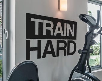 Puede incluir: Calcomanía de pared motivacional en blanco y negro con el texto "TRAIN HARD" en una pared blanca. Una máquina de ejercicio elíptica negra está en primer plano.