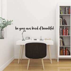 Pode incluir: Uma mesa branca com uma cadeira preta e uma l&acirc;mpada. A parede atr&aacute;s da mesa tem um adesivo de vinil preto que diz "be your own kind of beautiful".