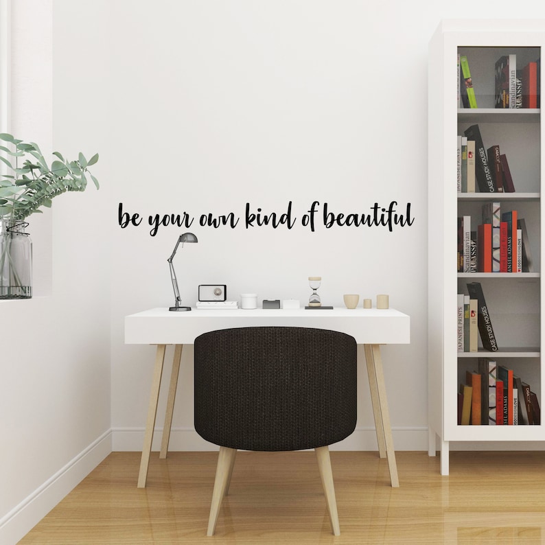 Pode incluir: Uma mesa branca com uma cadeira preta e uma l&acirc;mpada. A parede atr&aacute;s da mesa tem um adesivo de vinil preto que diz "be your own kind of beautiful".
