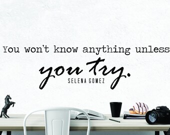May include: A white wall with a black and white inspirational quote that reads "You won't know anything unless you try." The quote is attributed to Selena Gomez. A white desk with a black horse figurine, a book, a black mug, a plate of doughnuts, and a black camera is in the foreground.