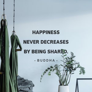 Puede incluir: Una cita motivacional en una pared blanca, "La felicidad nunca disminuye al compartirse. - Buda -" con una planta verde en una maceta blanca y un marco negro sobre una mesa en primer plano.