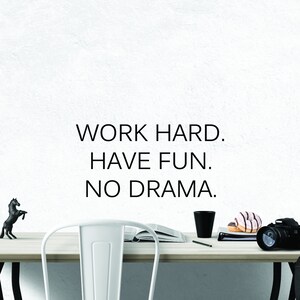 May include: A white desk with a black horse figurine, a book, a black mug, a pencil, a stack of books, two donuts, and a black camera. The wall behind the desk has the text "WORK HARD. HAVE FUN. NO DRAMA."