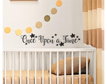 May include: White crib with a black vinyl decal that reads "Once Upon a Time" with stars on a white wall. A garland of yellow and gold circles hangs above the decal.