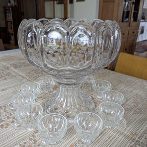 Op de afbeelding: Een heldere glazen punch bowl met een voet en een geschulpte rand, omringd door twaalf bijpassende kleine kopjes. De set wordt gepresenteerd op een tafelkleed met patroon, wat wijst op een vintage of antieke stijl, perfect voor het serveren van drankjes.