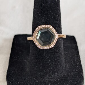 Pu&ograve; includere: Un anello in oro rosa con una gemma esagonale scura, circondata da un'aureola di piccole pietre trasparenti. L'anello &egrave; esposto su un espositore in velluto nero, che ne evidenzia l'elegante design e la lavorazione.