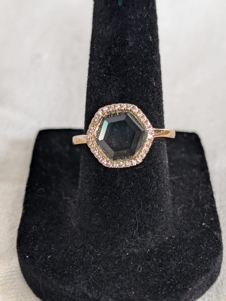 Pu&ograve; includere: Un anello in oro rosa con una gemma esagonale scura, circondata da un'aureola di piccole pietre trasparenti. L'anello &egrave; esposto su un espositore in velluto nero, che ne evidenzia l'elegante design e la lavorazione.