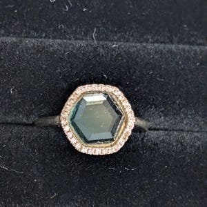 Pu&ograve; includere: Un anello d'argento con una gemma esagonale, verde scuro, circondata da un alone di piccole pietre trasparenti. L'anello &egrave; posto su uno sfondo di velluto nero, che mette in risalto la brillantezza delle gemme.