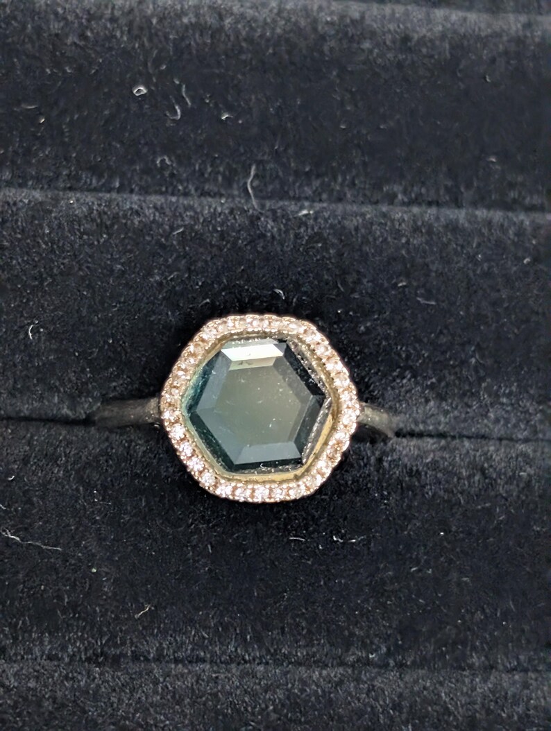 Pu&ograve; includere: Un anello d'argento con una gemma esagonale, verde scuro, circondata da un alone di piccole pietre trasparenti. L'anello &egrave; posto su uno sfondo di velluto nero, che mette in risalto la brillantezza delle gemme.