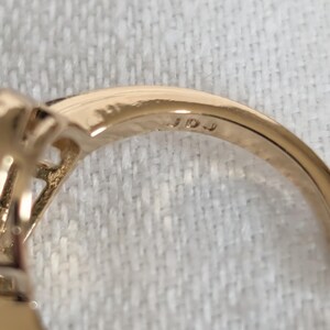 Pu&ograve; includere: Primo piano di un anello d'oro con incastonatura di pietre preziose. La fascia dell'anello &egrave; liscia e lucida, con le iniziali "JDJ" incise all'interno. L'anello &egrave; posto su uno sfondo di tessuto bianco.
