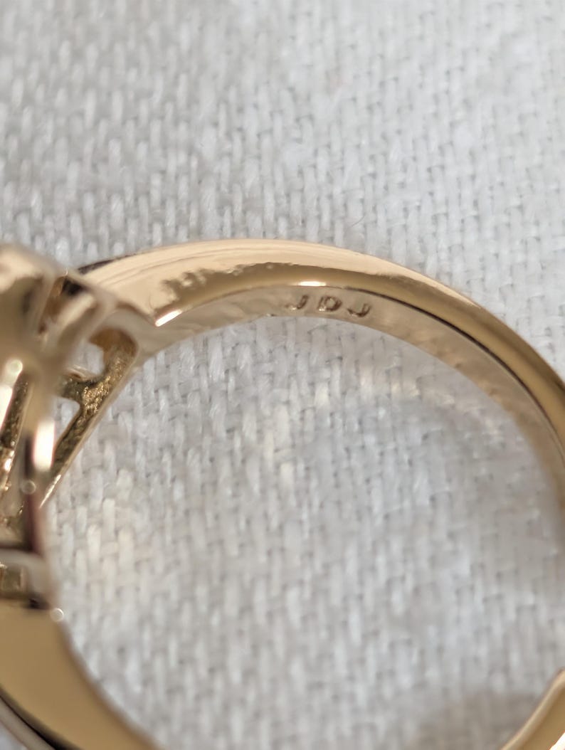 Pu&ograve; includere: Primo piano di un anello d'oro con incastonatura di pietre preziose. La fascia dell'anello &egrave; liscia e lucida, con le iniziali "JDJ" incise all'interno. L'anello &egrave; posto su uno sfondo di tessuto bianco.