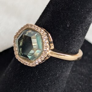 Pu&ograve; includere: Un anello d'oro con una grande pietra preziosa sfaccettata di colore verde acqua. La pietra preziosa &egrave; circondata da un alone di piccole pietre trasparenti. L'anello &egrave; presentato su una superficie nera, evidenziando il suo design elegante e la sua brillantezza.