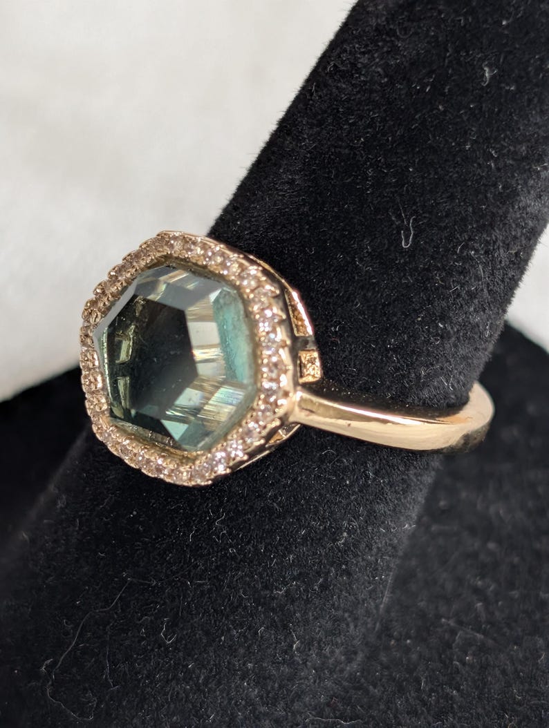 Pu&ograve; includere: Un anello d'oro con una grande pietra preziosa sfaccettata di colore verde acqua. La pietra preziosa &egrave; circondata da un alone di piccole pietre trasparenti. L'anello &egrave; presentato su una superficie nera, evidenziando il suo design elegante e la sua brillantezza.