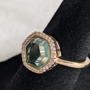 Pu&ograve; includere: Un anello d'oro con una grande gemma sfaccettata color teal, circondata da piccole pietre trasparenti. L'anello &egrave; appoggiato su una superficie nera, evidenziando i dettagli del gioiello.