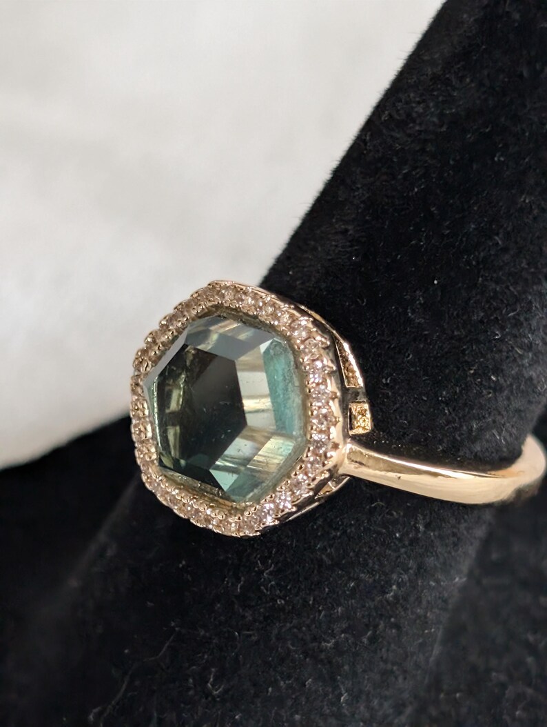 Pu&ograve; includere: Un anello d'oro con una grande gemma sfaccettata color teal, circondata da piccole pietre trasparenti. L'anello &egrave; appoggiato su una superficie nera, evidenziando i dettagli del gioiello.