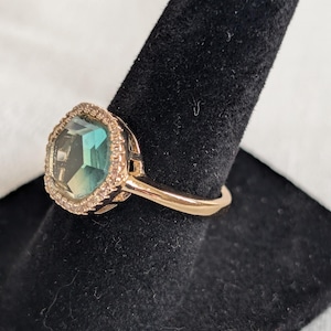 Pu&ograve; includere: Un anello dorato con una grande pietra preziosa di colore verde acqua, tagliata a forma quadrata. La pietra preziosa &egrave; circondata da un alone di piccole pietre trasparenti. L'anello &egrave; esposto su un supporto nero.