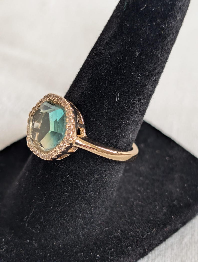 Pu&ograve; includere: Un anello dorato con una grande pietra preziosa di colore verde acqua, tagliata a forma quadrata. La pietra preziosa &egrave; circondata da un alone di piccole pietre trasparenti. L'anello &egrave; esposto su un supporto nero.