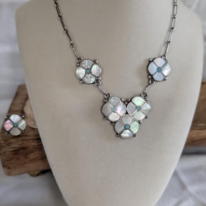 May include: A silver-toned necklace and matching ring set featuring flower-shaped motifs. Each flower is composed of four iridescent mother-of-pearl petals surrounding a small, round, bright blue center stone set in a silver-colored metal frame.