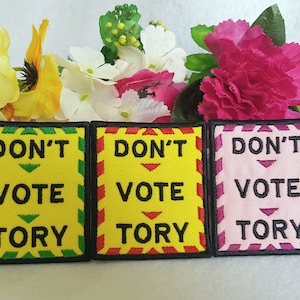 May include: Three embroidered patches with the text "DON'T VOTE TORY". The patches are yellow, red, and pink, with green and black borders. The patches are set against a backdrop of colourful flowers.