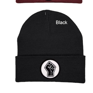 Pode incluir: Quatro gorros de malha em diferentes cores: bordô, cinza, preto e verde-garrafa. Cada gorro tem um círculo branco com um punho preto em relevo no centro.