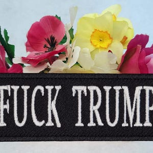 May include: A black embroidered patch with the words "FUCK TRUMP" in white, set against a backdrop of colorful flowers including pink, yellow, and white blooms. The patch is rectangular.