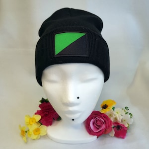 May include: Black beanie with a green and black patch on the front. The beanie is on a white mannequin head with flowers around the base.
