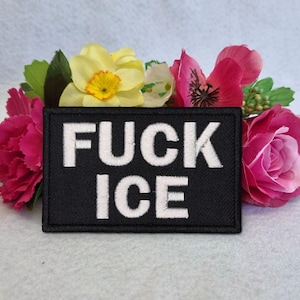 Pode incluir: Um patch preto com as palavras "FUCK ICE" em letras brancas, rodeado por flores artificiais rosa, amarelas e verdes. As flores incluem rosas, cravos e outras flores.
