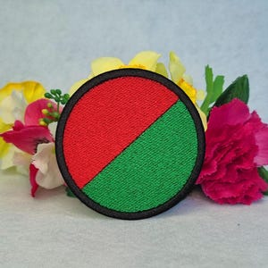 May include: A circular embroidered patch with a black border, split diagonally into red and green segments. The patch is displayed with a backdrop of colourful flowers, including yellow, pink, and white blooms.