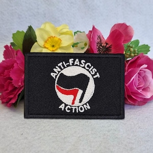 May include: A rectangular black embroidered patch featuring a circular white emblem with a black and red flag design in the center. The text ANTI-FASCIST ACTION is stitched in white thread, curving around the top and bottom of the central circle.