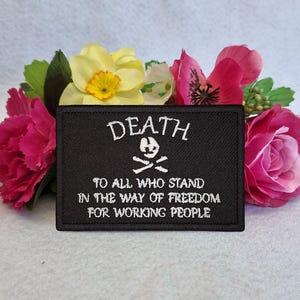 Może przedstawiać: Prostokątna czarna haftowana naszywka z białym napisem DEATH TO ALL WHO STAND IN THE WAY OF FREEDOM FOR WORKING PEOPLE oraz małą białą ikoną czaszki z piszczelami umieszczoną centralnie pomiędzy słowem DEATH a resztą tekstu.