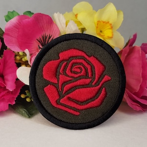 Może przedstawiać: Naszywkę z haftowaną czerwoną różą na ciemnozielonym tle z czarną obwódką. Naszywka jest otoczona różowymi i żółtymi kwiatami.