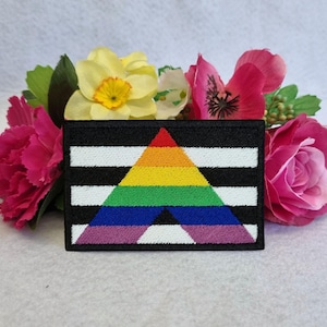 Op de afbeelding: Een zwart-wit gestreept patch met een regenboogdriehoek in het midden. De driehoek is gemaakt van de kleuren rood, oranje, geel, groen, blauw, indigo en violet. De patch is omringd door roze bloemen.