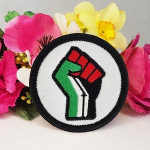 May include: A black and white circular patch with a raised, green, red, and black fist in the center. The fist is raised in the air with the index finger pointing up.