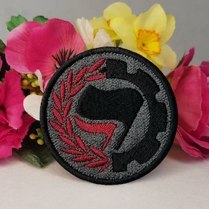 May include: A black and gray embroidered patch with a red stylized fist and a wreath around it. The patch is circular and has a gear design on the right side.